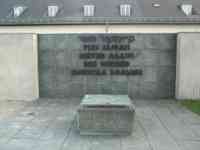 Square tomb in front of a wall marked "Never Again" in five languages